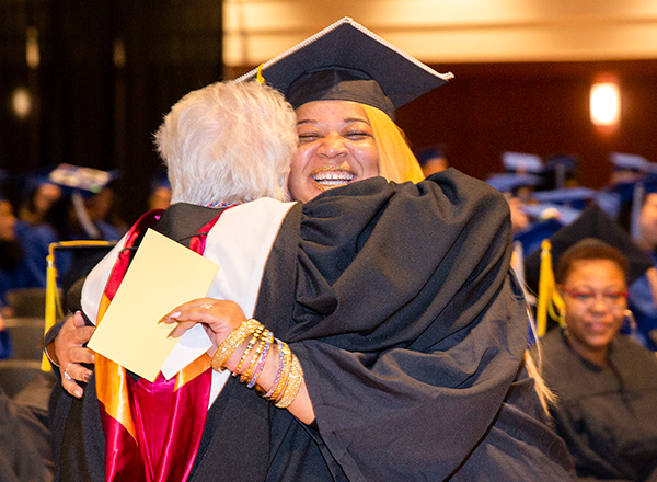 Smiling graduate with degree in hand hugs professor in auditorium at commencement.